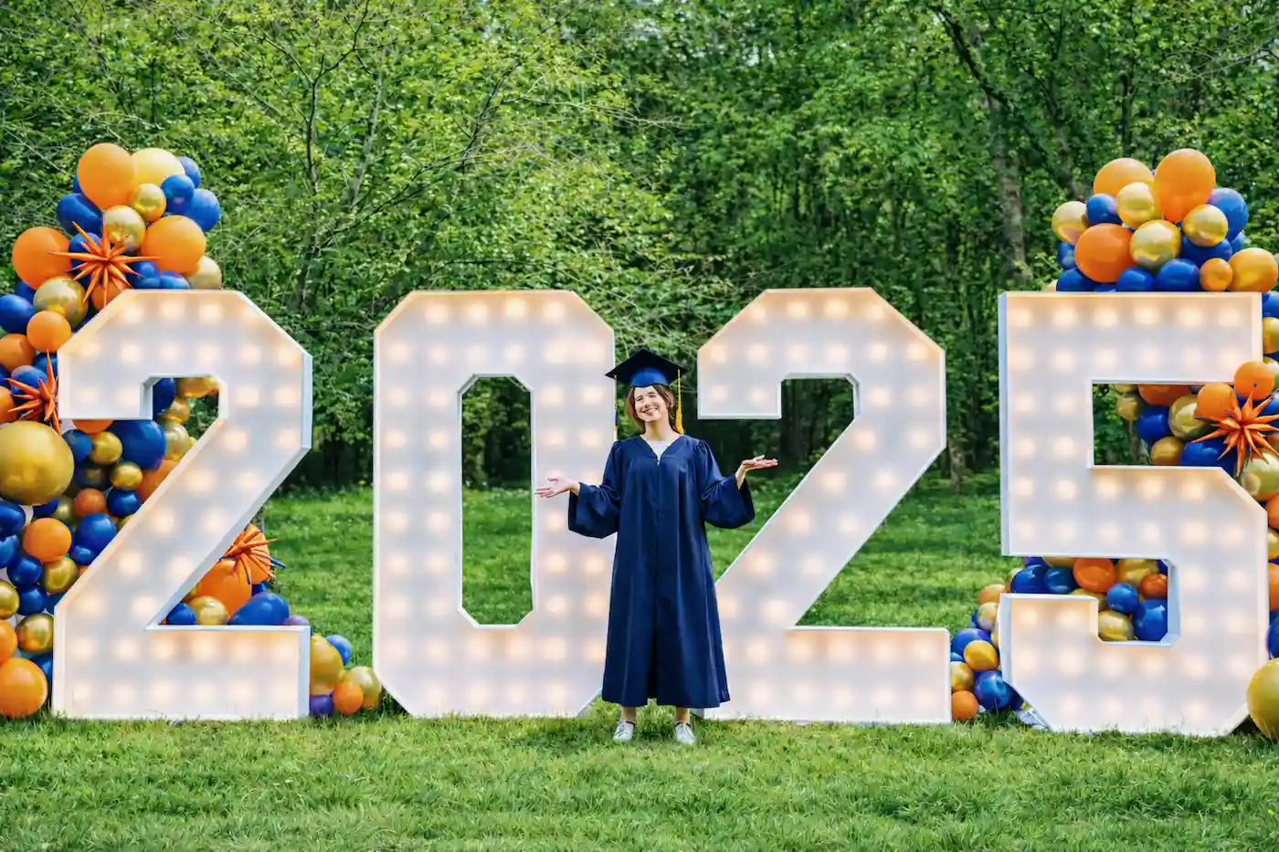 Graduate standing in front of light-up 2025 marquee letters with balloons - grad letters