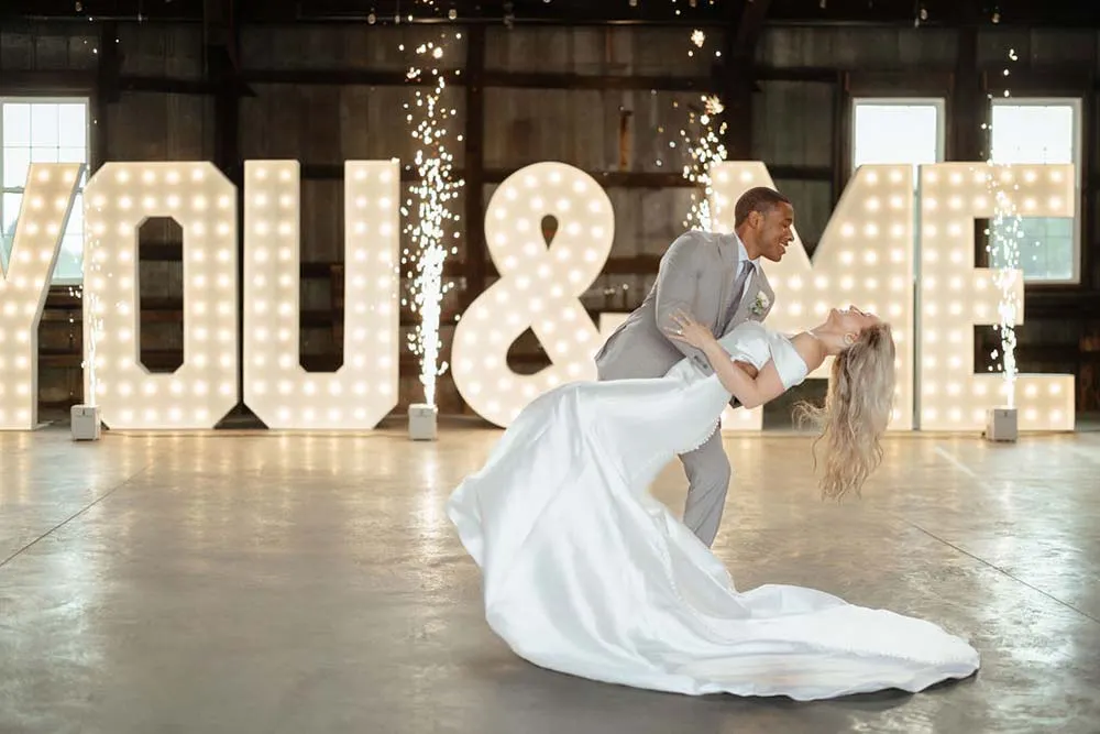 Bride and groom dip kiss in front of YOU & ME marquee letters with sparklers