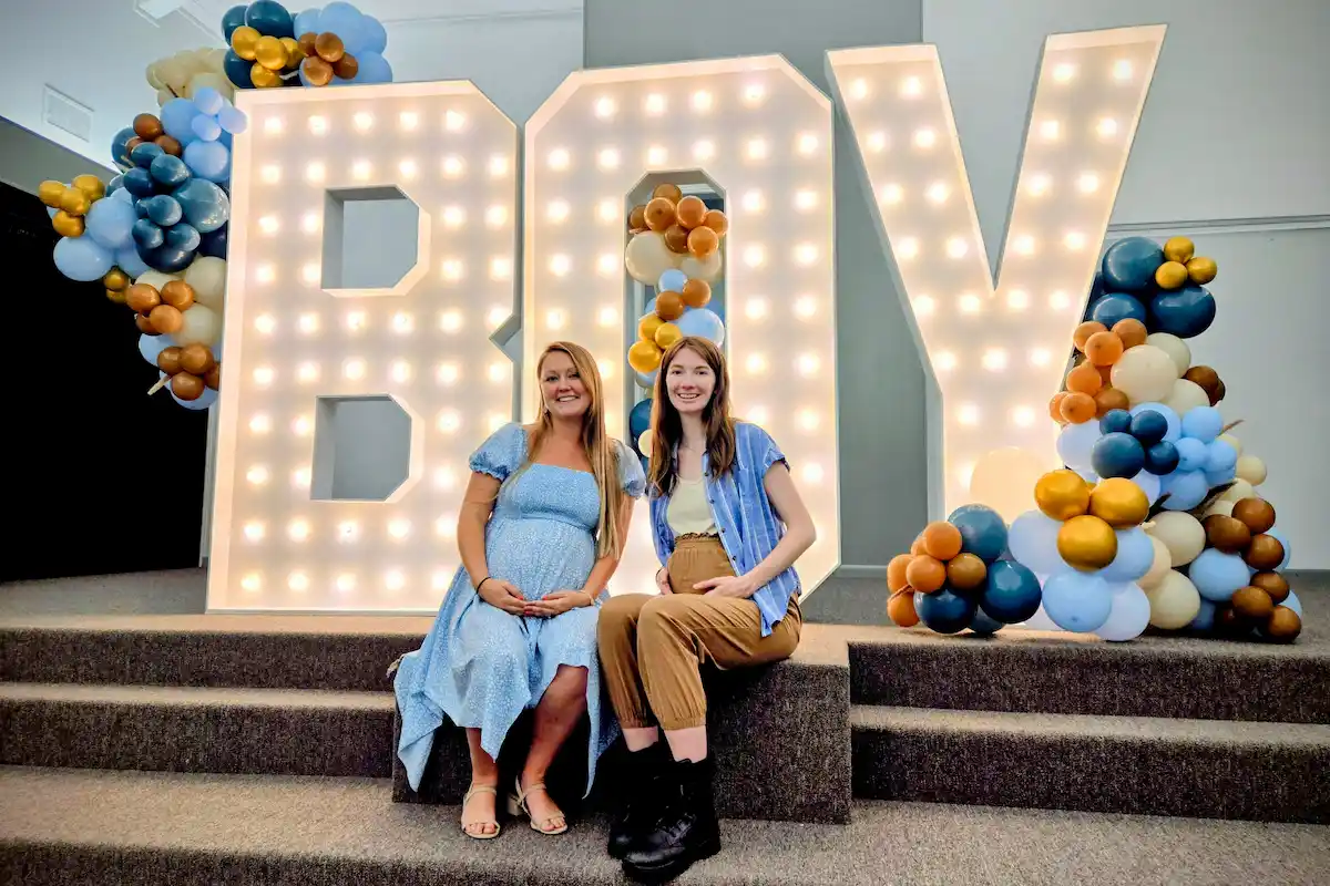 Two women standing in front of BOY gender reveal marquee letters with balloons
