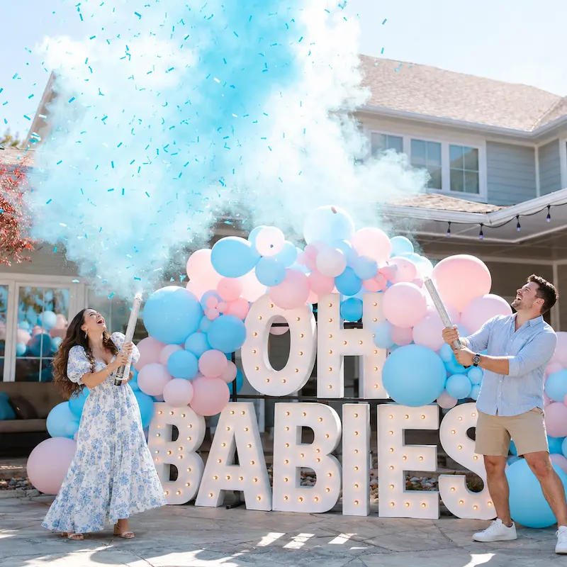 Couple celebrating with blue smoke in front of stacked OH BABIES marquee letters and pink and blue balloons