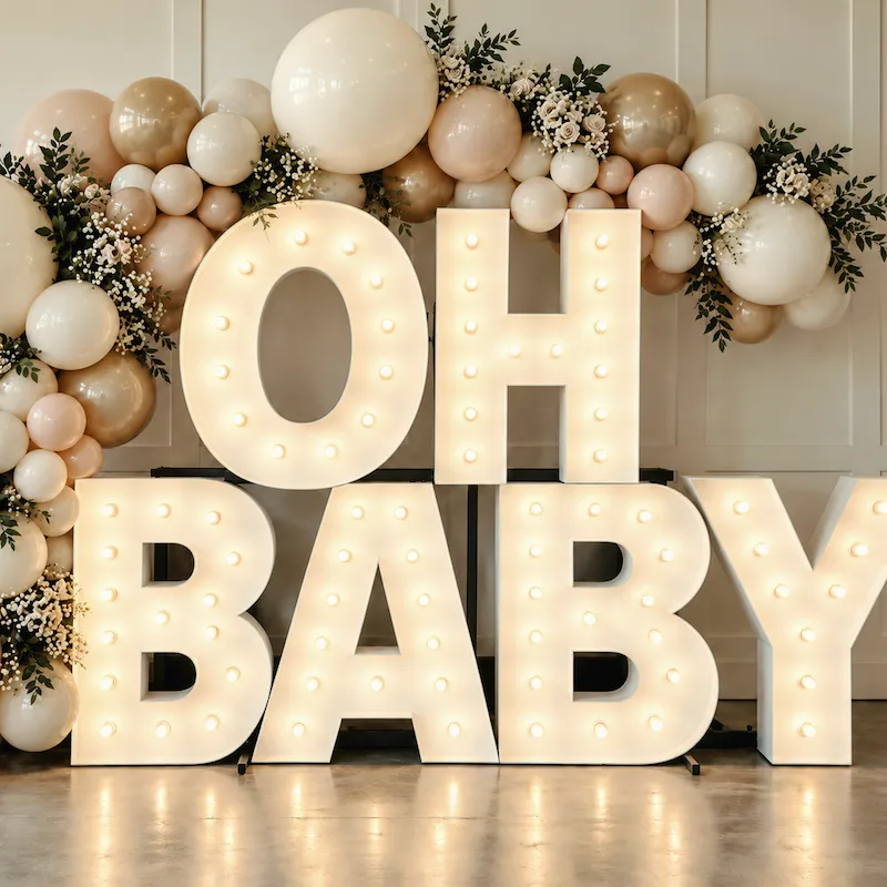 Prizes for baby shower games displayed beside stacked OH BABY light-up letters with blush, ivory and champagne balloons